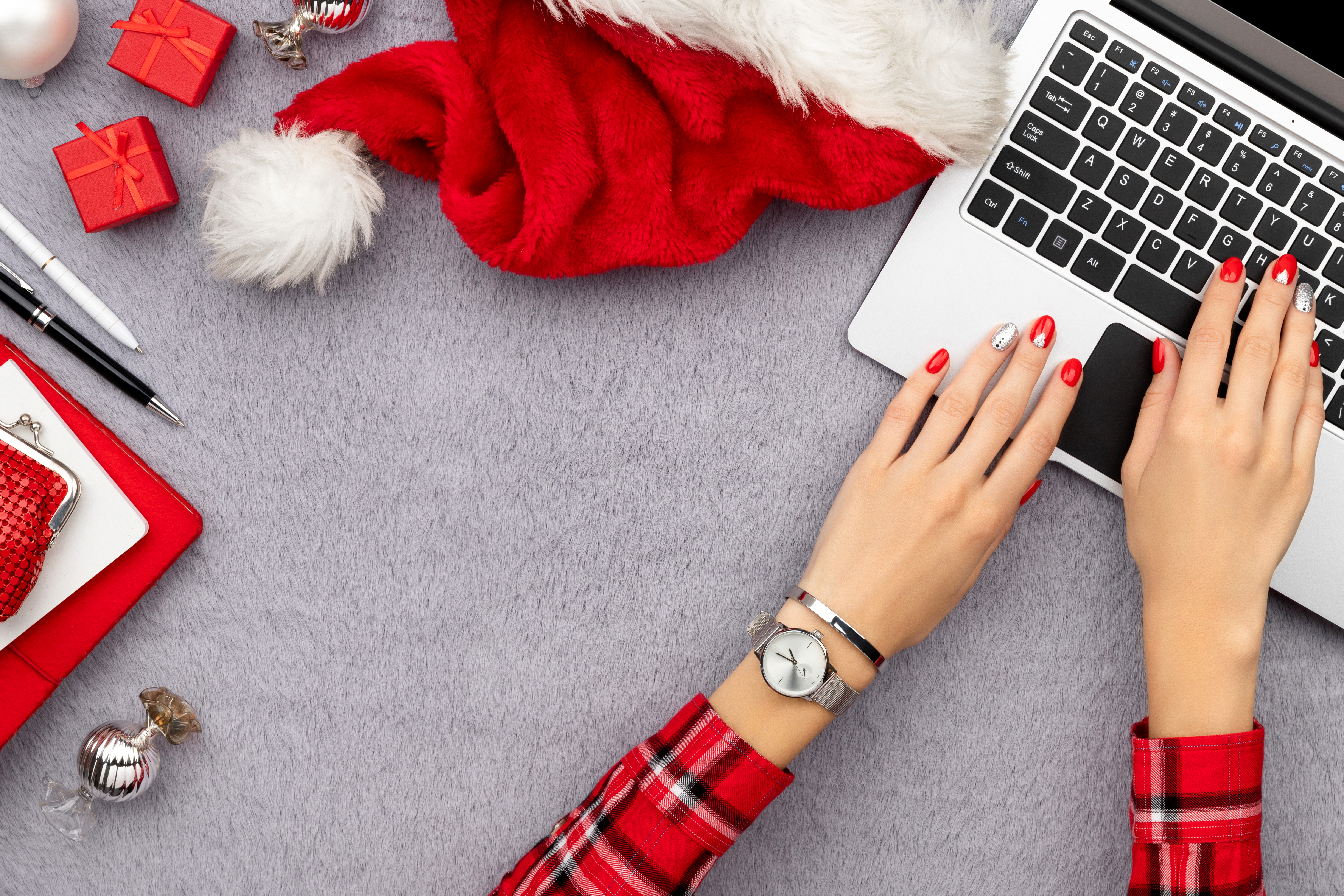 Eine Frau in rot-karierten Oberteil tippt auf einem Laptop. Sie hat rot und silber lackierte Nägel und trägt eine silberne Armbanduhr und ein silbernes Armband. Neben dem Laptop liegen eine rote Weihnachtsmütze, kleine rote Geschenke, silberner Tannenbaumschmuck, ein rotes Notizbuch, ein roter Geldbeutel, weiße Karten und ein schwarzer Kugelschreiber.